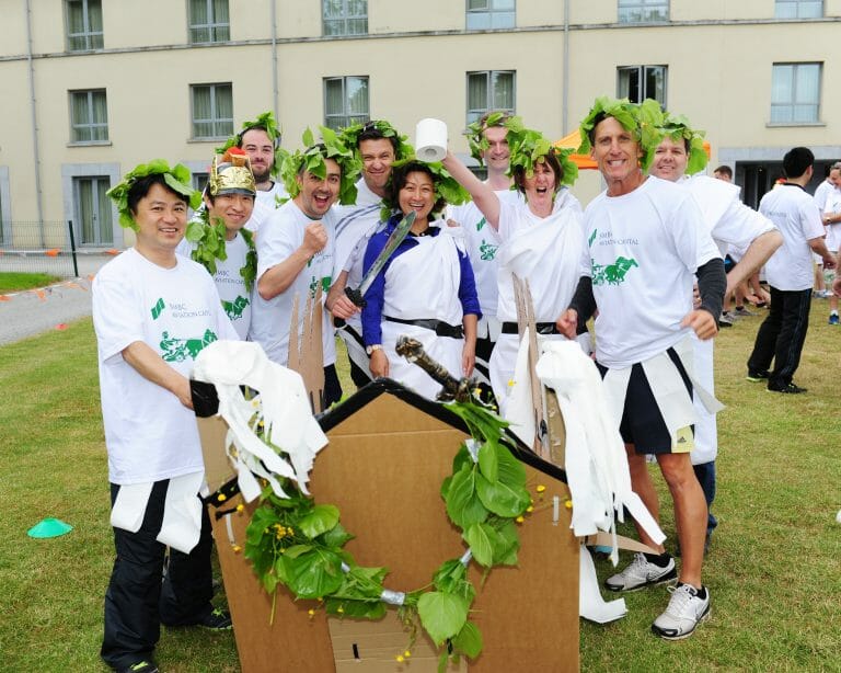 Delegates dressed in roman clothing standing next to the cardboard chariot they built during their corporate away day.