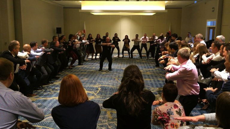 Group of delegates standing in a circle, while Ed, an Orangeworks instructor teaches them how to do the Haka