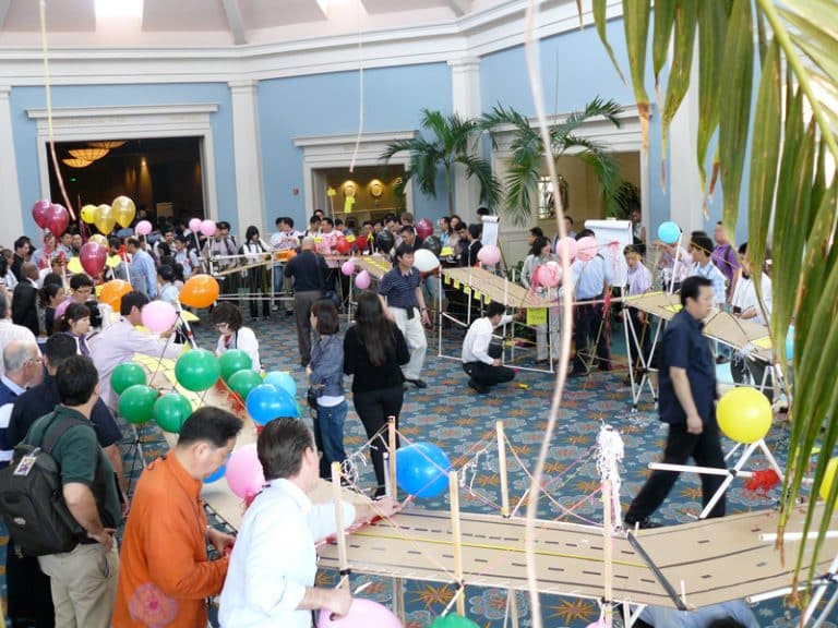 Teams stand around their completed bridge they have constructed, during a fun team building experience with Orangeworks.