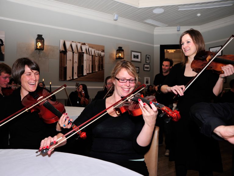 3 delegates smiling and laughing while learning how to play the violin, as part of their staff away day with Orangeworks.