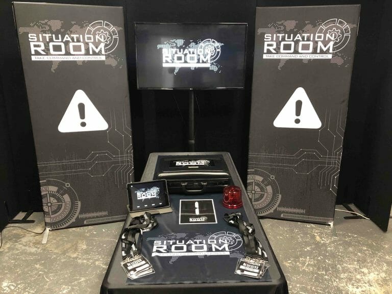 the situation room with equipment laid out on the table, including a briefcase, an iPad and lanyards with a tv in the back.