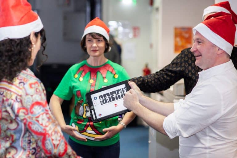A man holding an ipad showing it to his colleague while another lady stands behind them wearing a santa hat, during their festive team building challenge with Orangeworks.