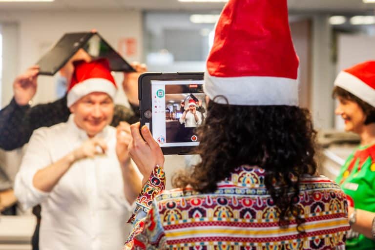 A woman holding an ipad taking a photo of a wearing a Santa hat with a book on his head, as part of their Christmas team building activity with Orangeworks.
