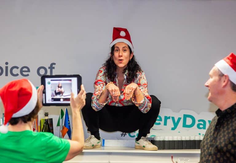 A man holding an ipad taking a photo of a woman doing a monkey pose on a table while wearing a Santa hat, as part of their Christmas themed team building activity with Orangeworks.