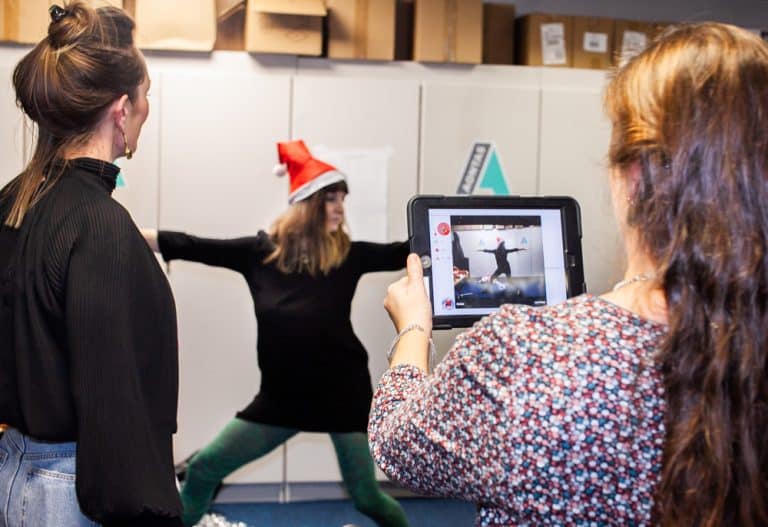 A lady holding an ipad taking a photo of a woman doing a yoga pose in a Santa hat, as part of their Christmas themed team building activity with Orangeworks.
